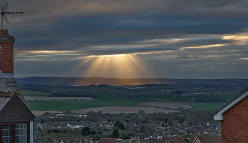 This landscape photograph captures a rural scene in Derbyshire, United Kingdom, during a spring evening as witnessed from the hillside houses of Bolsover in England. The main feature of the image is the striking display of God's rays, also known as crepuscular rays, streaming dramatically through breaks in the clouds and illuminating the distant landscape. Below this natural phenomenon, a solar farm with clearly visible rows of solar panels stretches across the fields, highlighting the blend of natural beauty and renewable energy within the region. The houses in the foreground emphasize the rural community setting, while the rolling hills and patchwork fields of Derbyshire serve as a picturesque backdrop enhanced by the soft, diffused light of the spring evening.